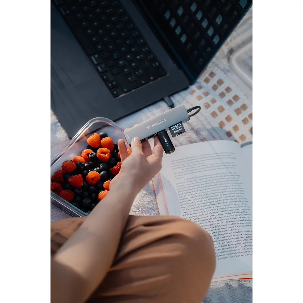 Person hält einen grauen USB-C-Hub mit eingesteckten SD-Karten neben einem Laptop, Beeren und einem aufgeschlagenen Buch.