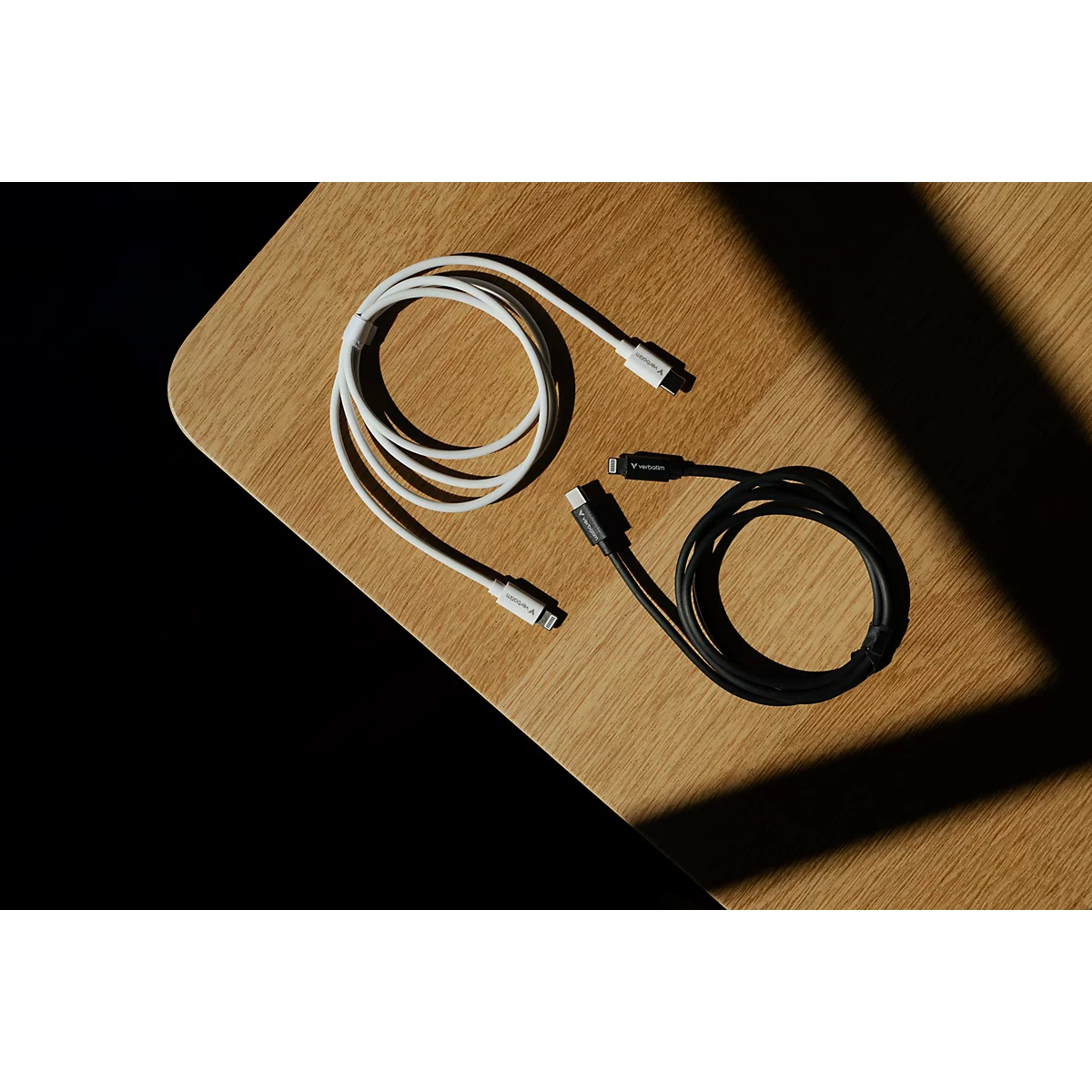 Ein weißes und ein schwarzes Ladekabel liegen in Schlaufen auf einem hölzernen Tisch im Licht und Schatten.