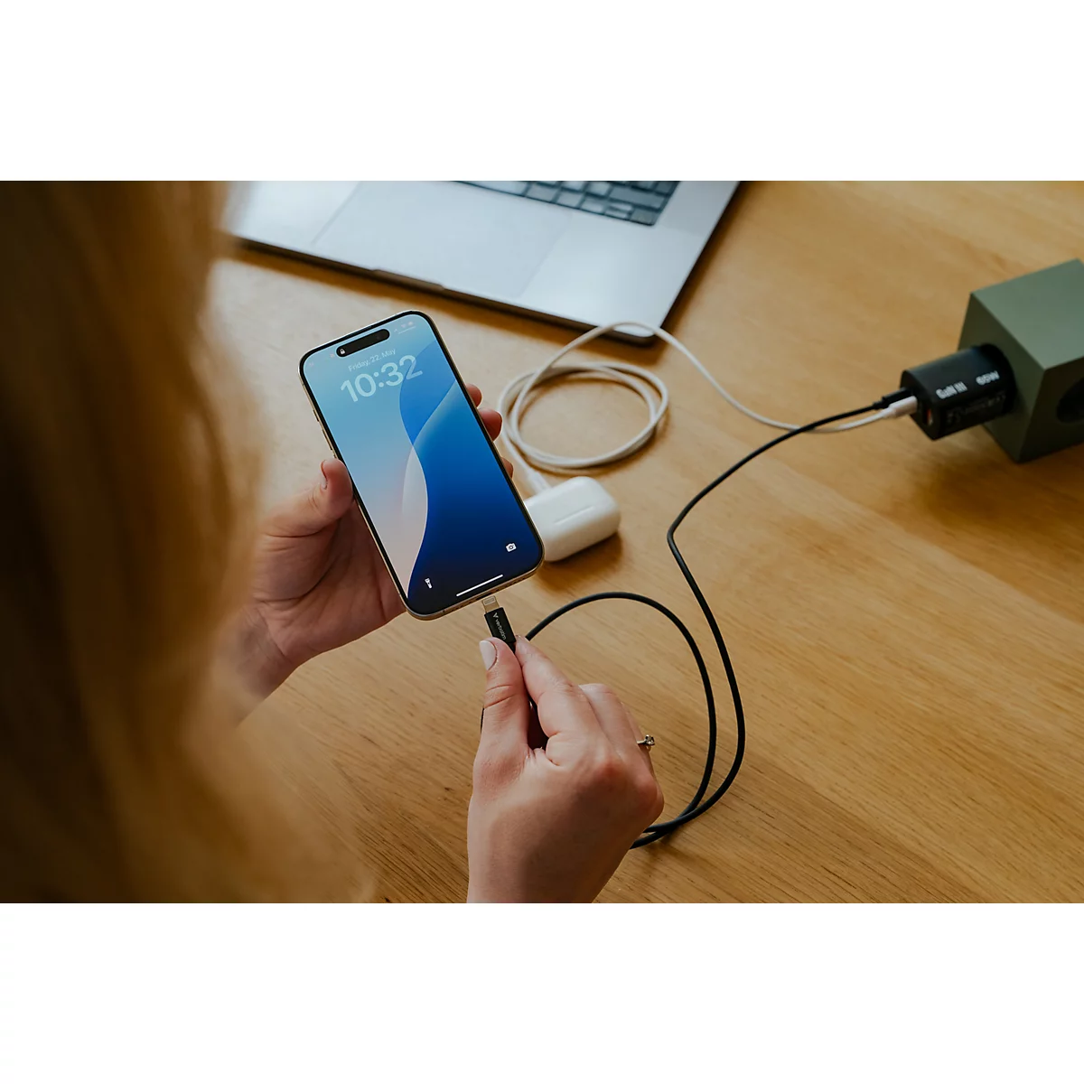 Person hält ein Smartphone, das an einem Kabel mit einem Ladestecker an einem Holztisch verbunden ist.