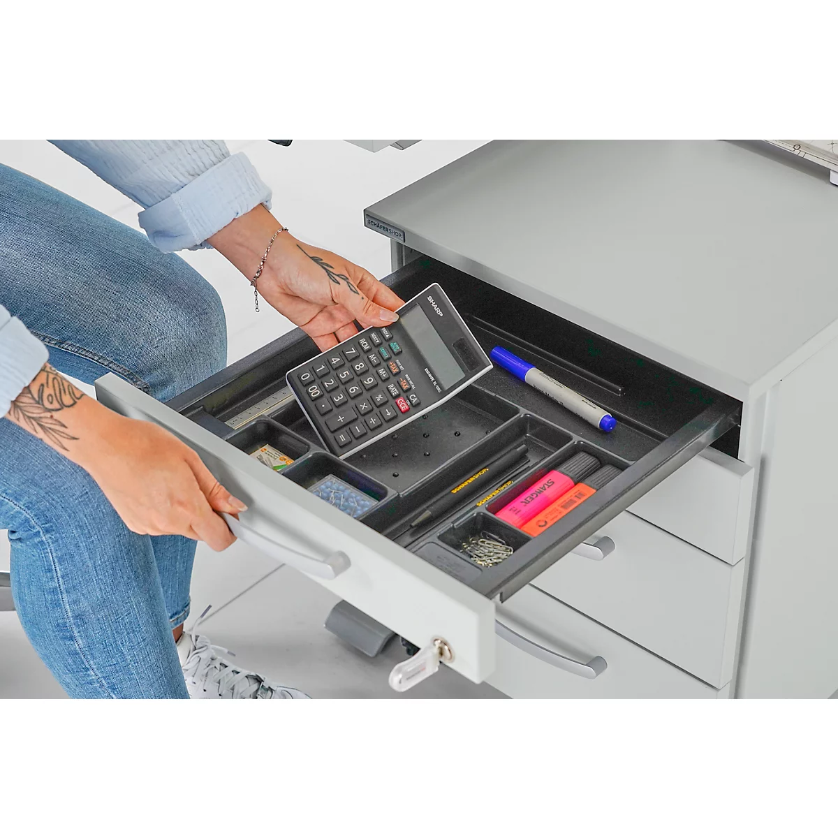 Persoon opent een lade van een kantoorkast met pennen, rekenmachine, markeerstiften en paperclips.