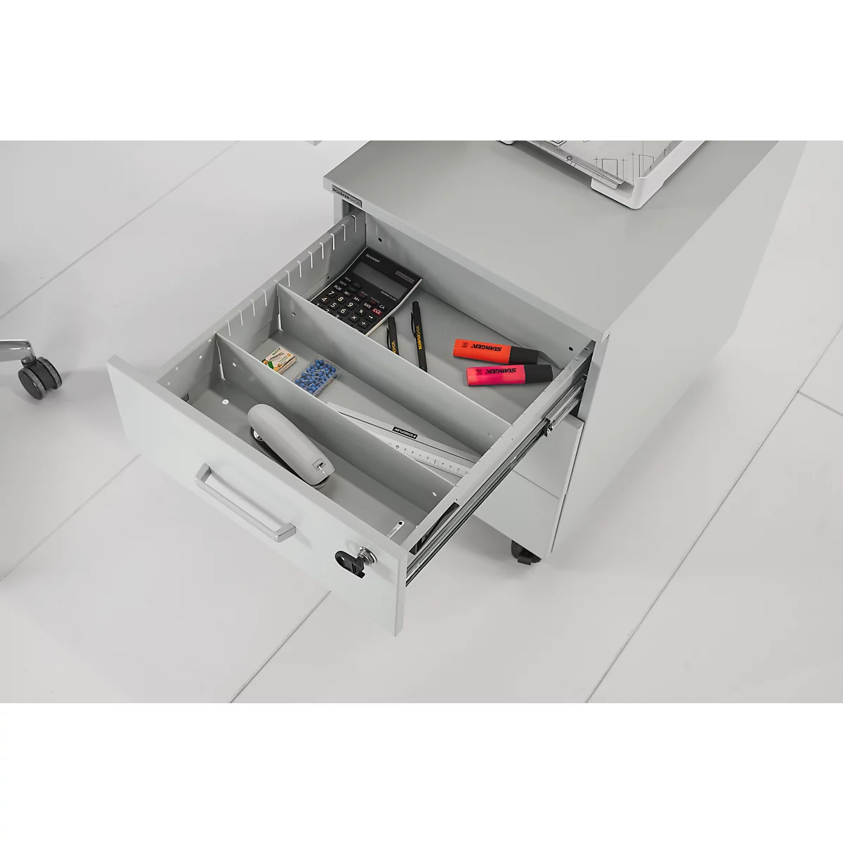Grijze rolcontainer met een open lade. Het bevat een rekenmachine, pennen, markeerstiften, een liniaal en andere kantoorbenodigdheden.