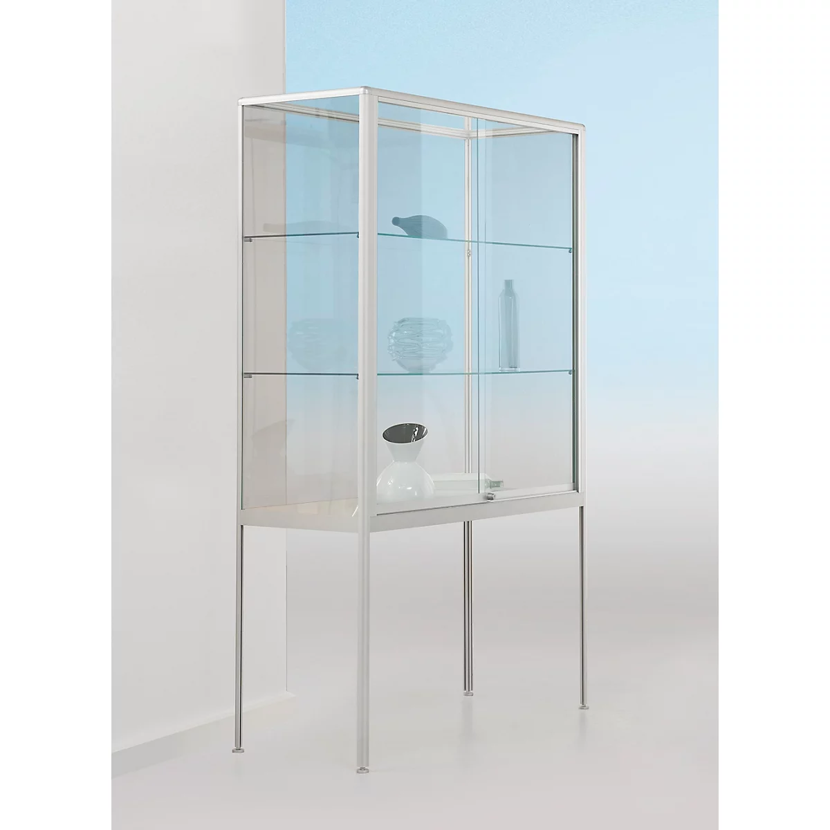 Un meuble vitrine en verre avec un cadre en métal se tient devant un mur bleu clair. À l'intérieur, des objets comme une bouteille noire et une sculpture blanche sont exposés.