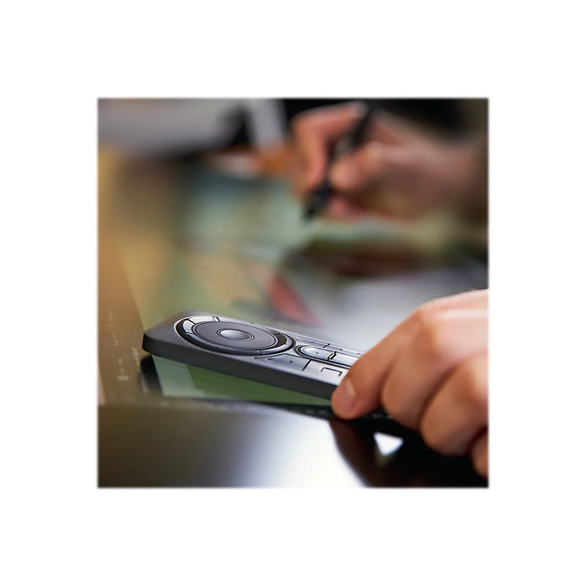 Nahaufnahme einer Hand, die eine schwarze Fernbedienung über einem Touchscreen hält. Im Hintergrund wird mit einem Stift gearbeitet.