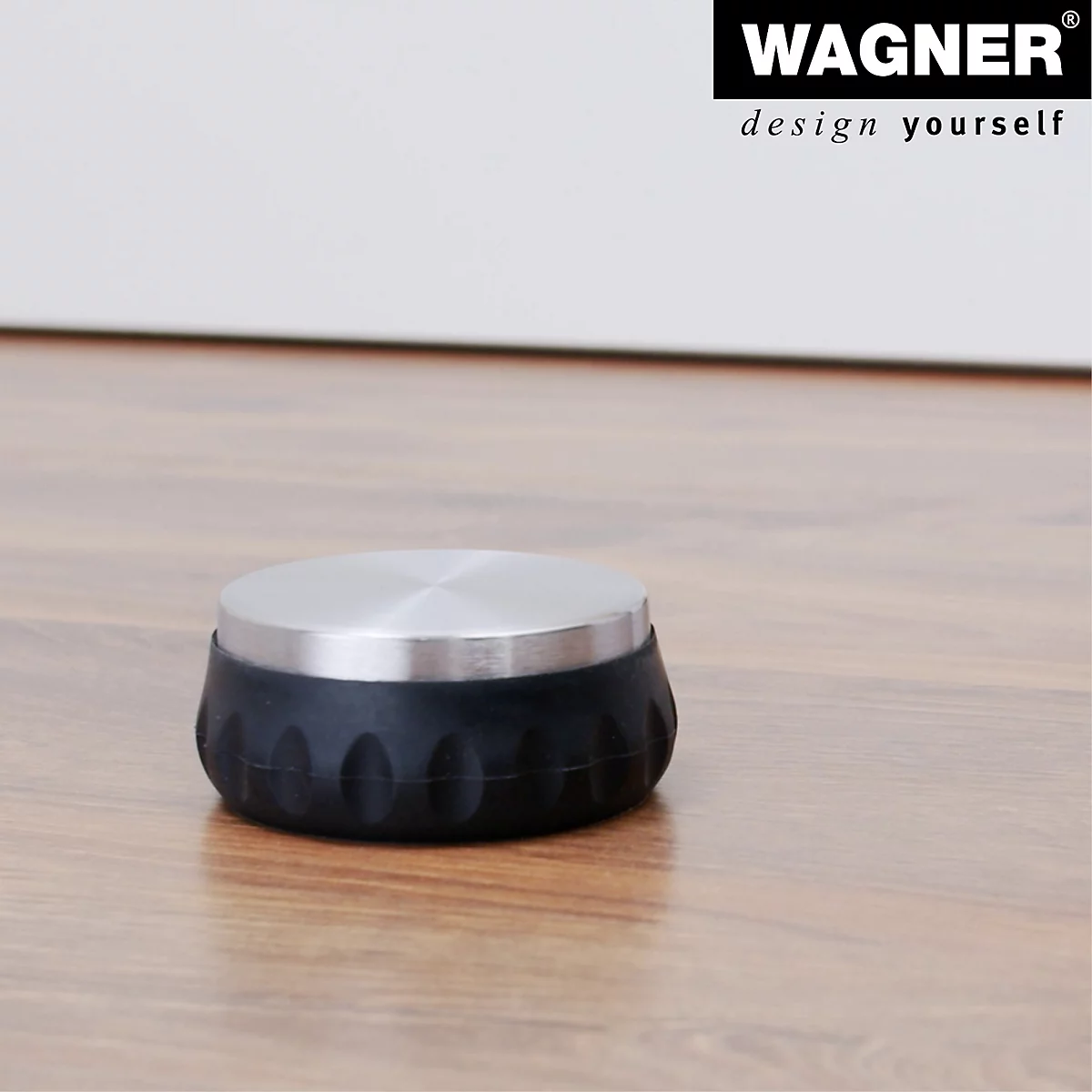 Ein schwarzer Türstopper mit silberner Oberseite liegt auf einem Holzboden. Darüber Logo "WAGNER design yourself".