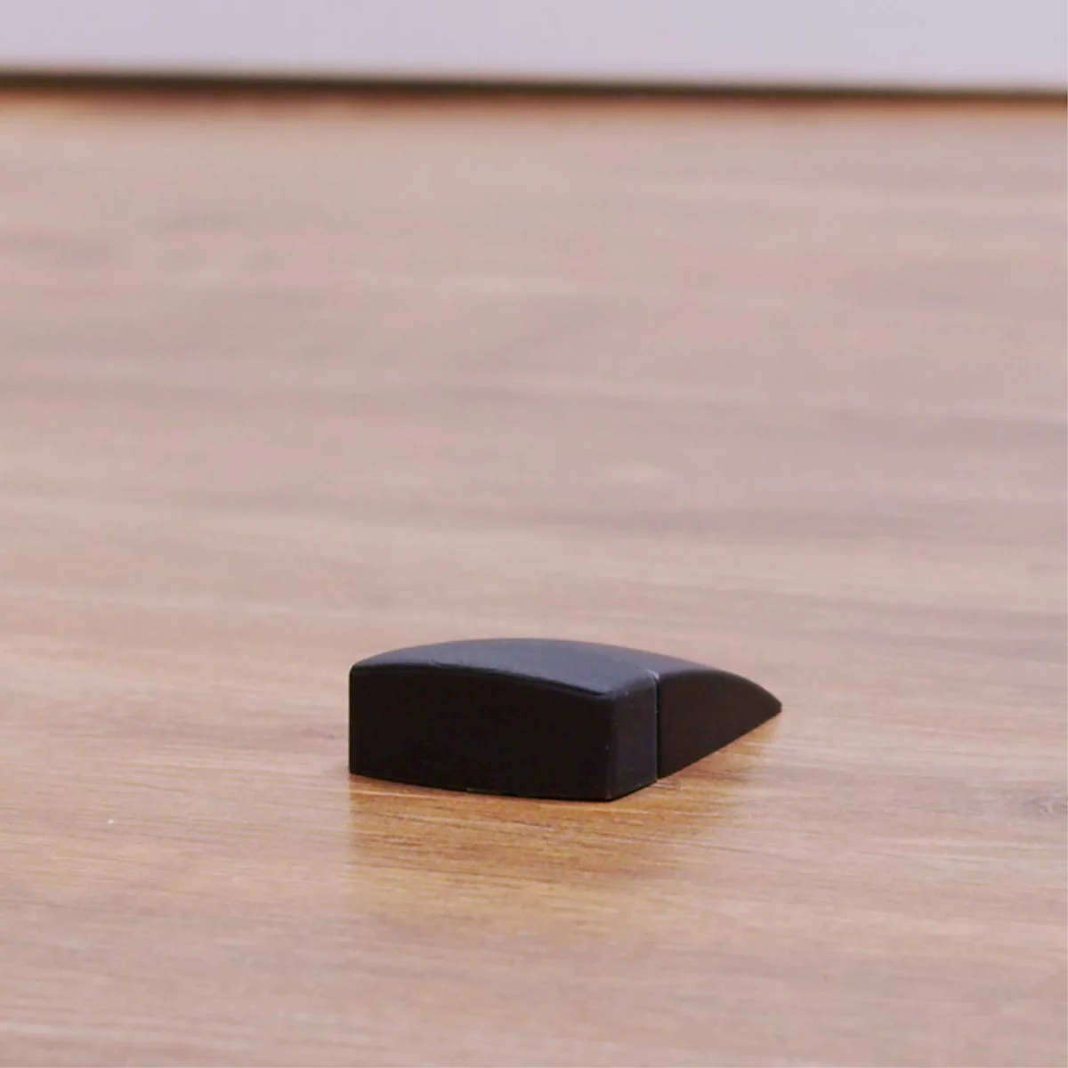 Een zwarte houten deurstopper ligt op een lichtbruine houten vloer. De stopper heeft een rechthoekige vorm met een schuine kant.
