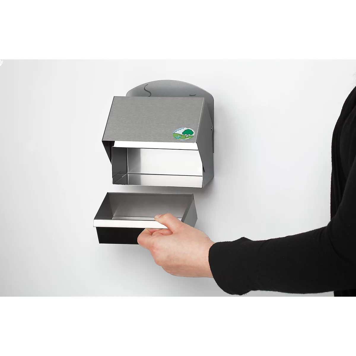 A person opens a metal paper towel dispenser on a white wall. The dispenser has a green logo.