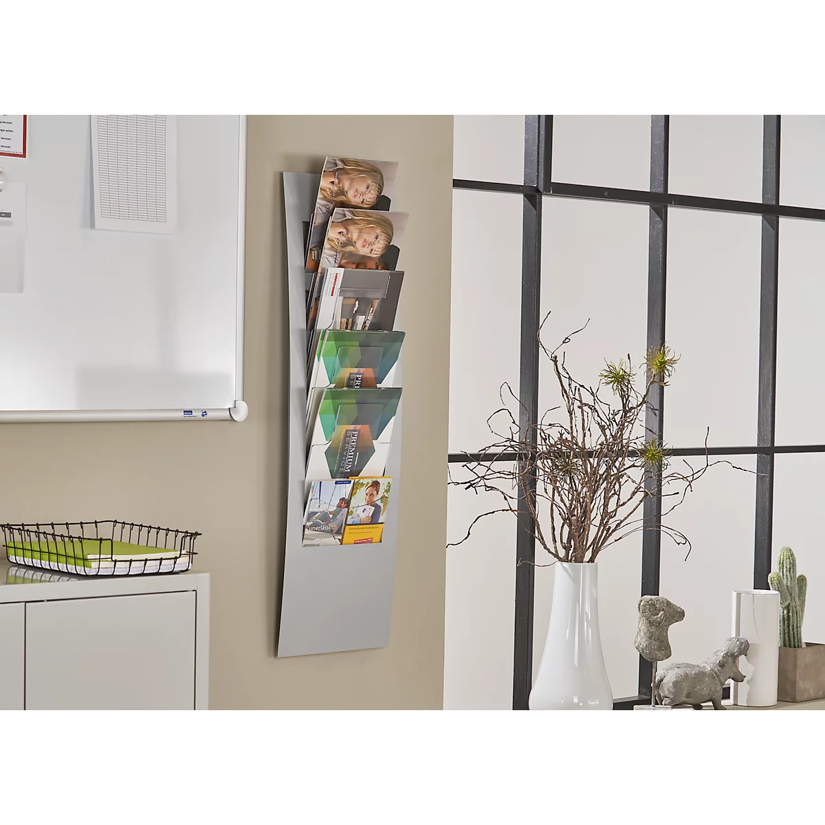 Grey wall newspaper with brochures, next to it window, flower vase and decoration.