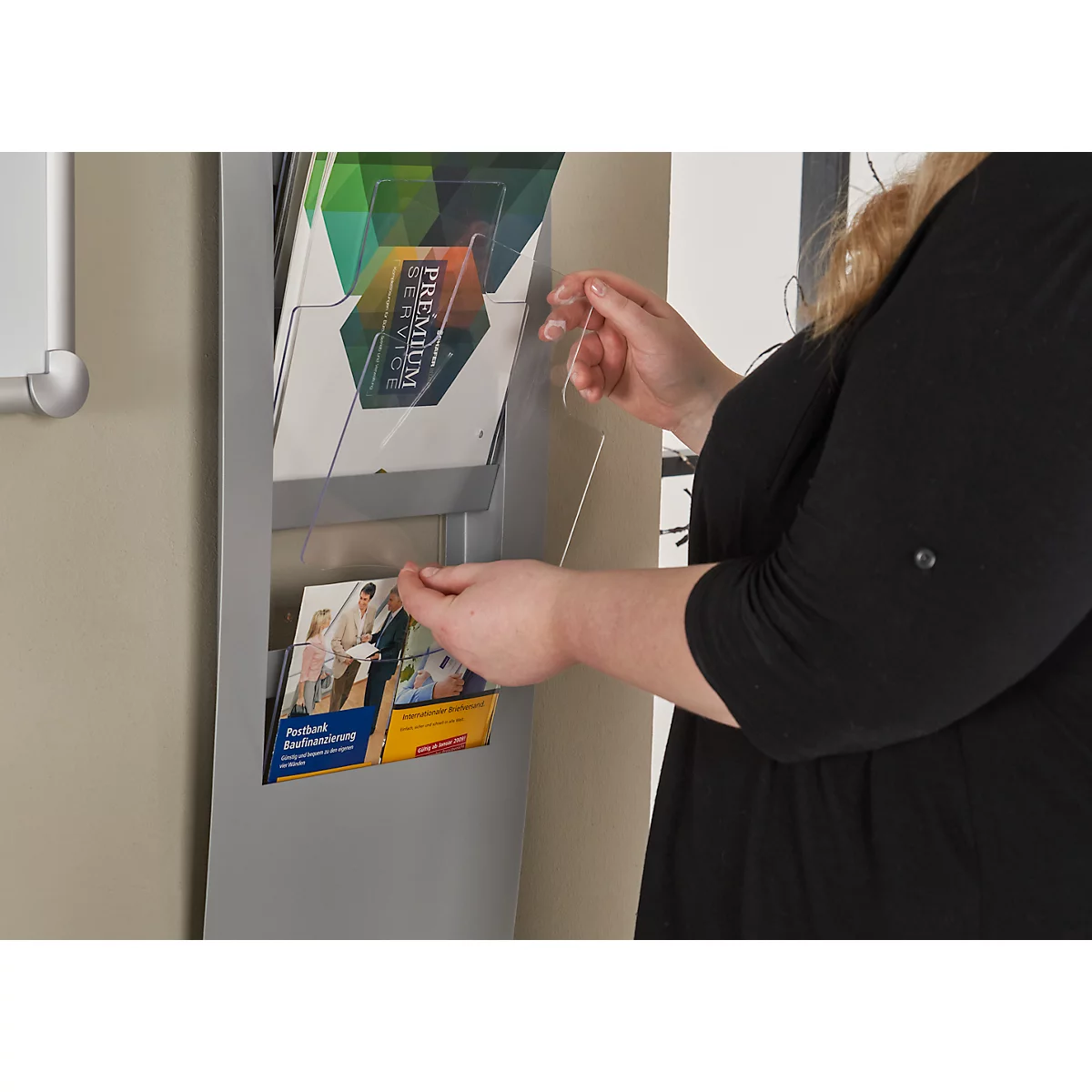A woman opens a brochure compartment. Brochures with prints can be seen inside. The woman is wearing a black top.