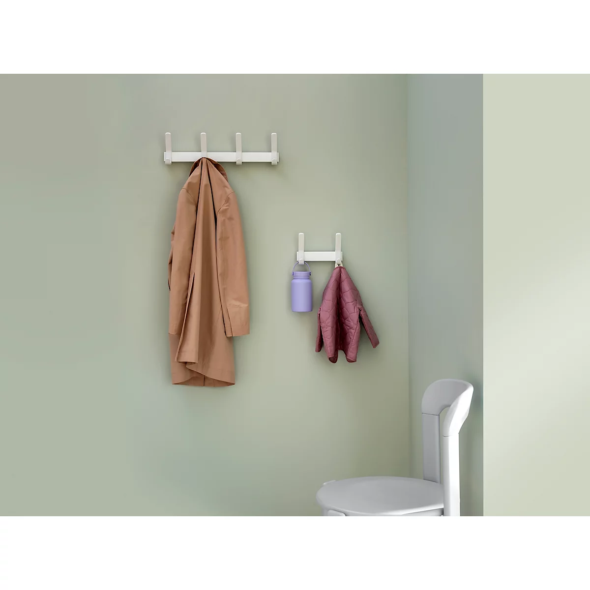 Zwei weisse Garderobenhaken an einer Wand. An dem oberen hängt ein brauner Mantel, an dem unteren eine rote Jacke und eine lila Flasche.