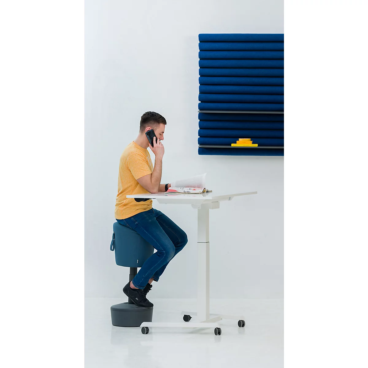 Man aan de telefoon aan zijn bureau. Hij zit op een ergonomische stoel. Blauw akoestisch paneel aan de muur.