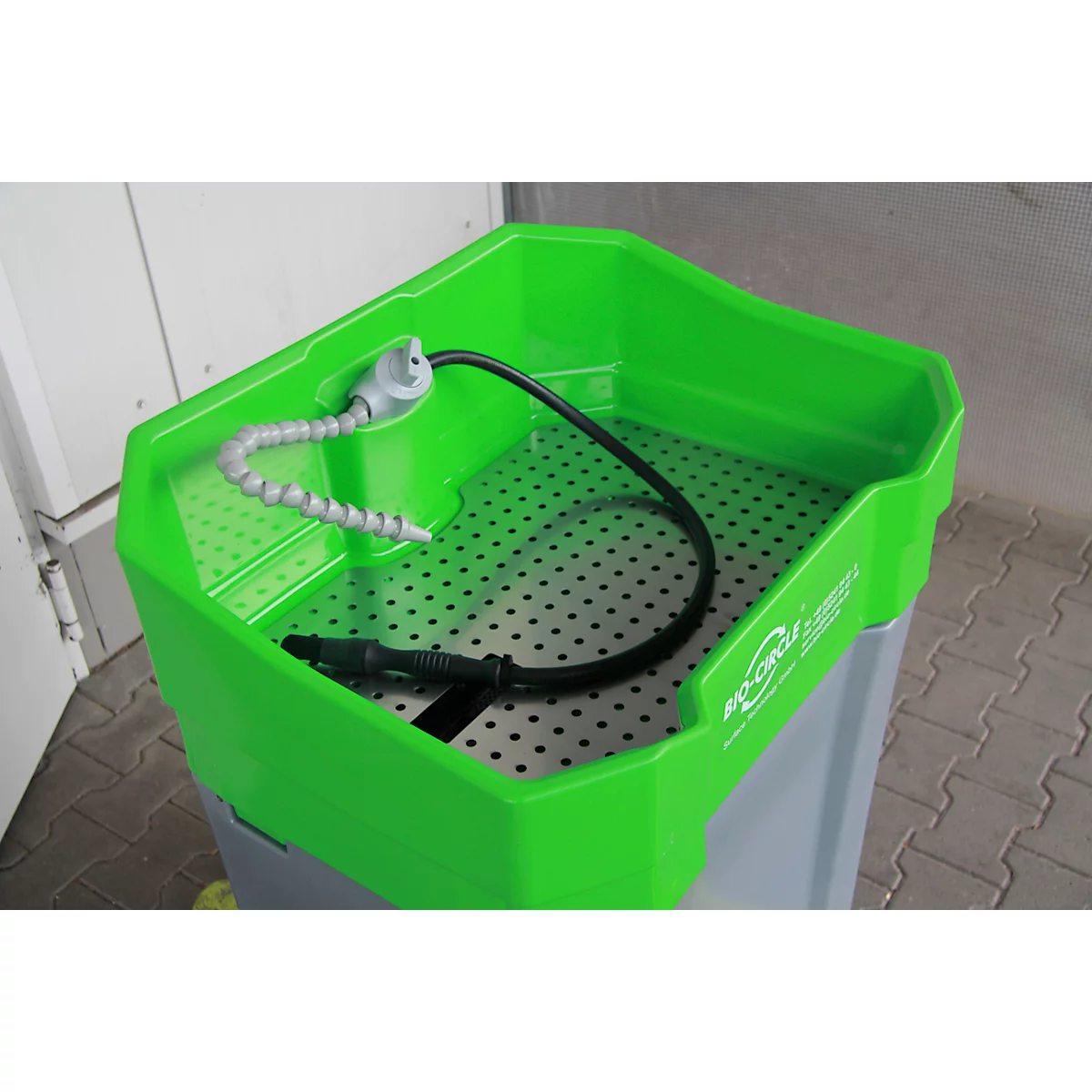 Grüne Waschbox mit integrierter Halterung für einen Wasserschlauch. Der Behälter steht auf einem grauen Unterbau.