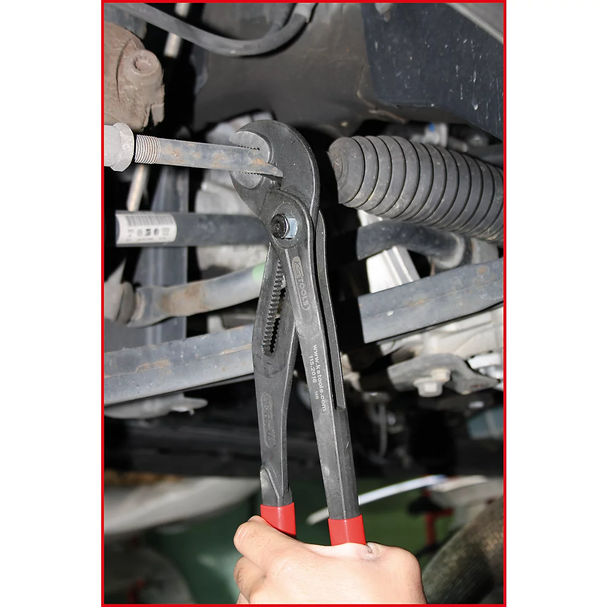 Eine Person hält eine Zange an einem Autoteil. Roter Griff, graue Zange, Autoteile im Hintergrund.