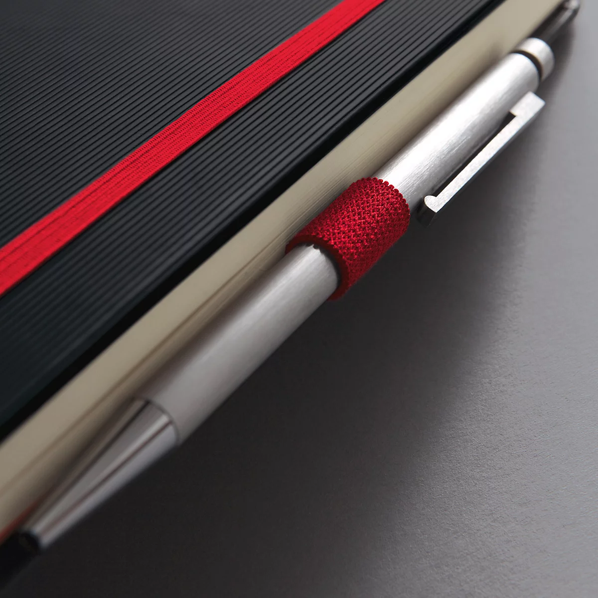 Close-up van een notitieboek met een rode elastiek en pen. De pen heeft een rode grip en een zilveren punt.