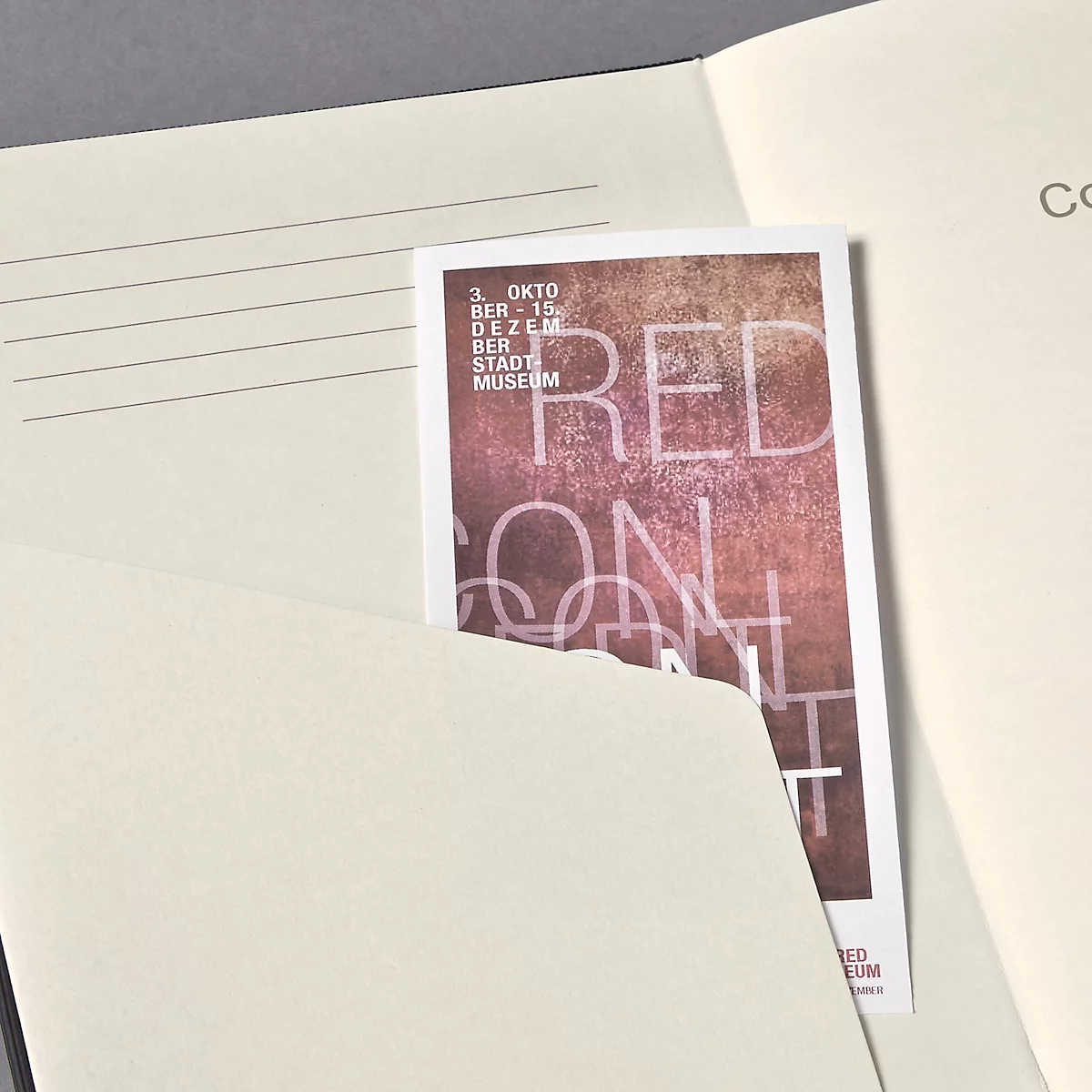 Een rode flyer in een notitieboek. De flyer toont in grote letters "RED CON..." en de evenementdata: 3. OKT - 15. DEC. BER STADTMUSEUM.