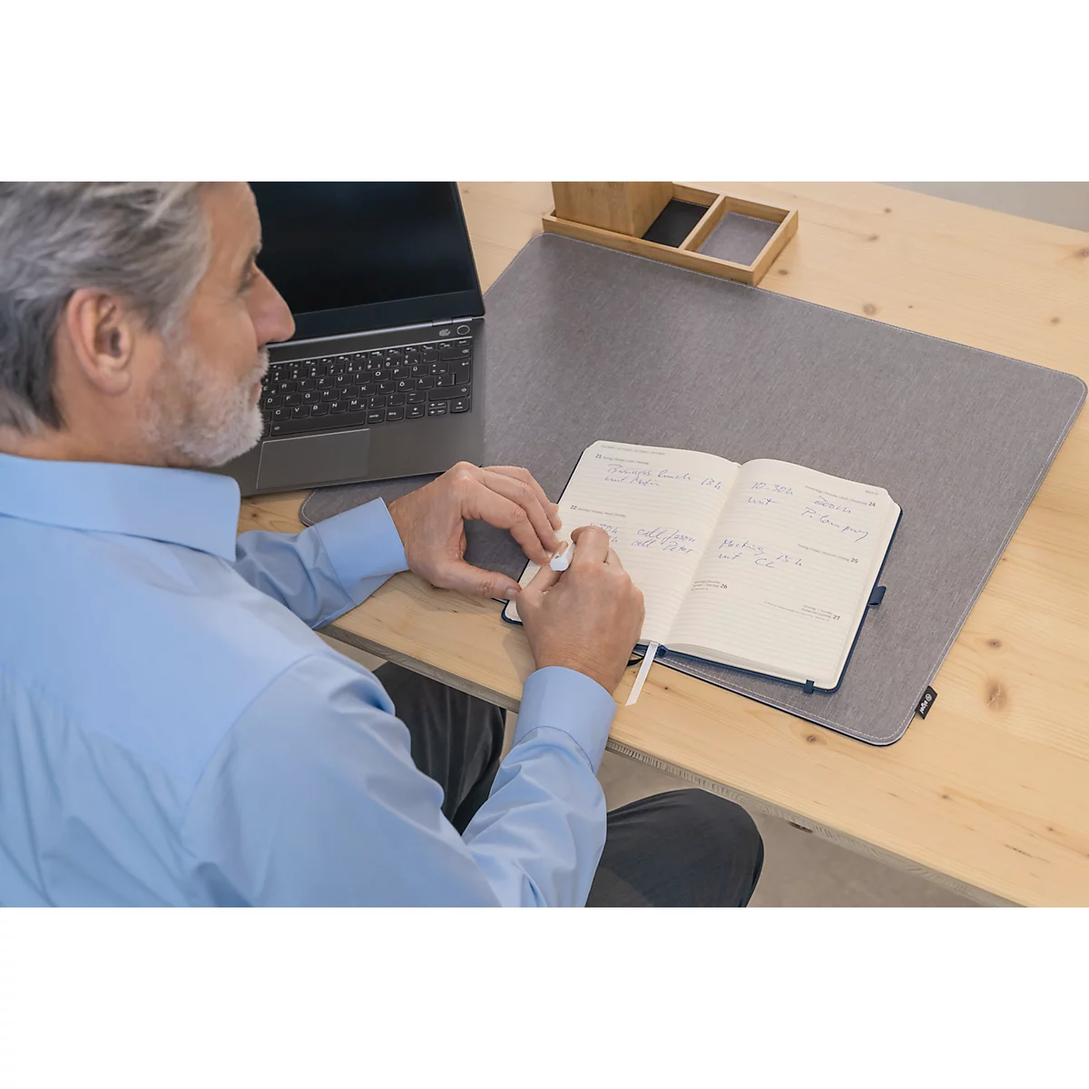 Een man schrijft in een notitieboekje op een bureau. Op de tafel liggen een grijze bureaublad en een laptop.