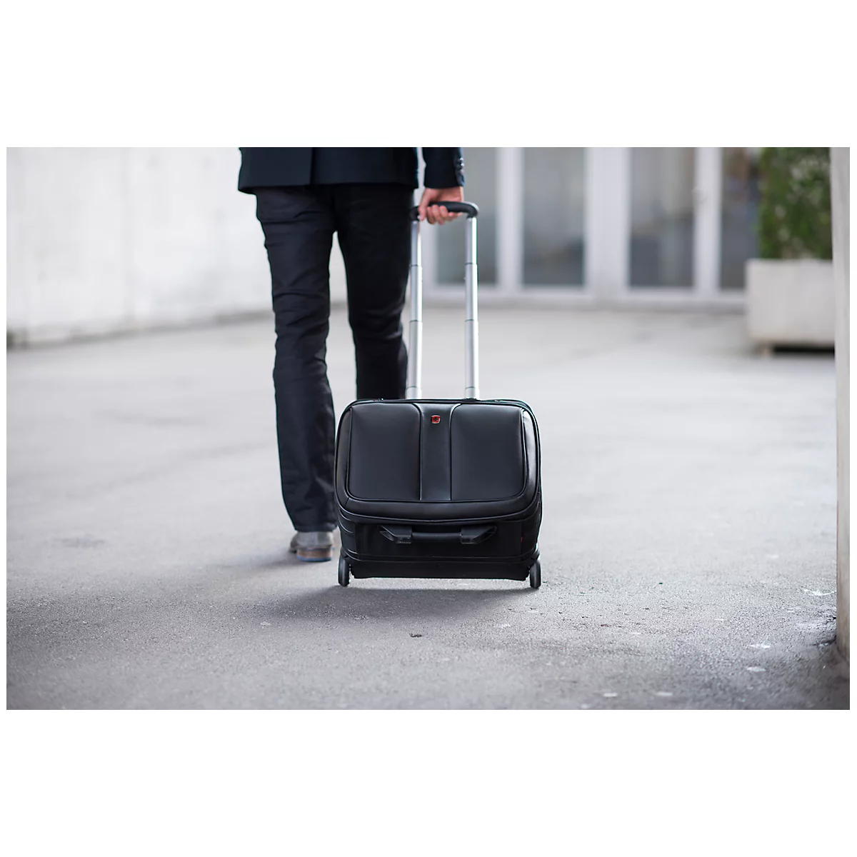 Person pulls a black trolley behind them, walking down a bright hallway.