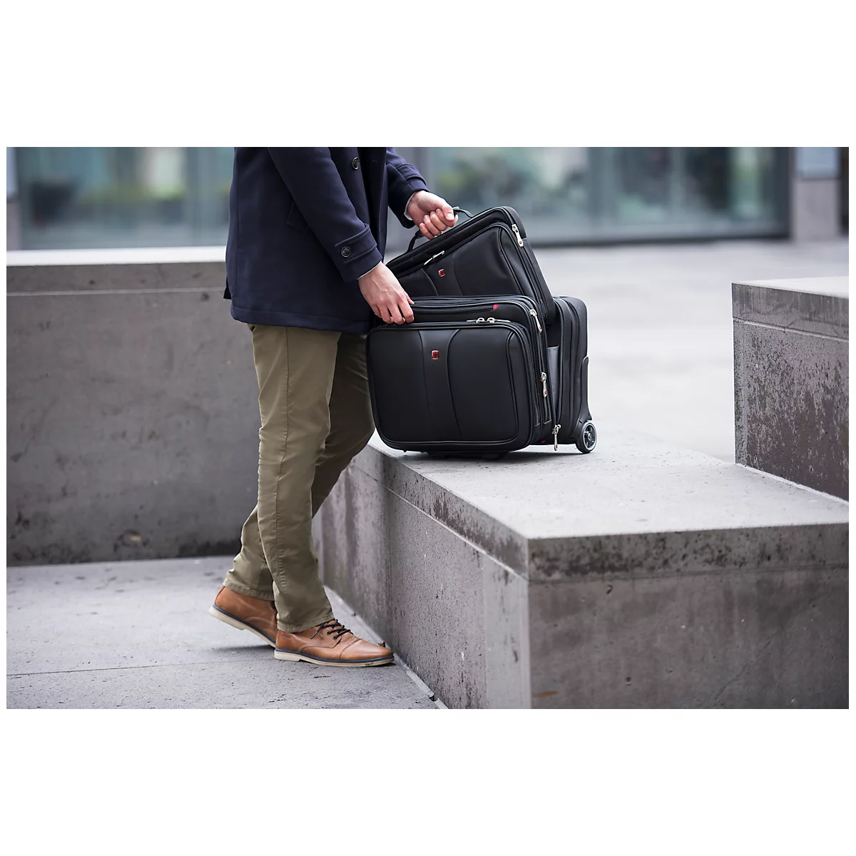 A person in olive pants and brown shoes carries several pieces of black luggage. The background is blurred.