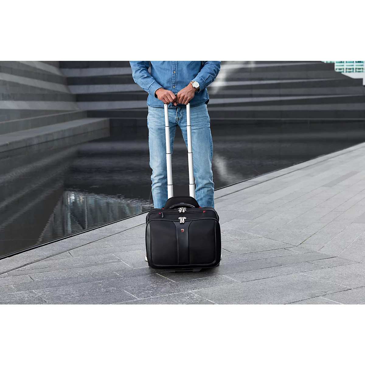 Man standing with a rolling suitcase. He is wearing jeans and a denim shirt.
