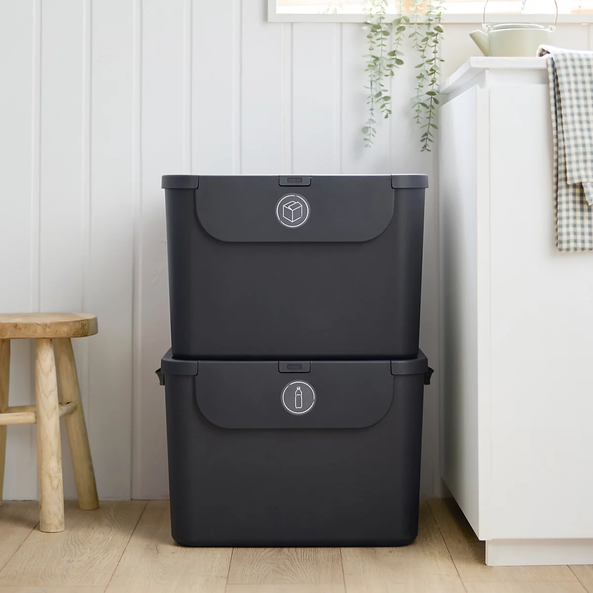 Person wirft Flasche in schwarzen Müllbehälter. Der Behälter steht auf einem weissen Mülleimer in einer Küche.