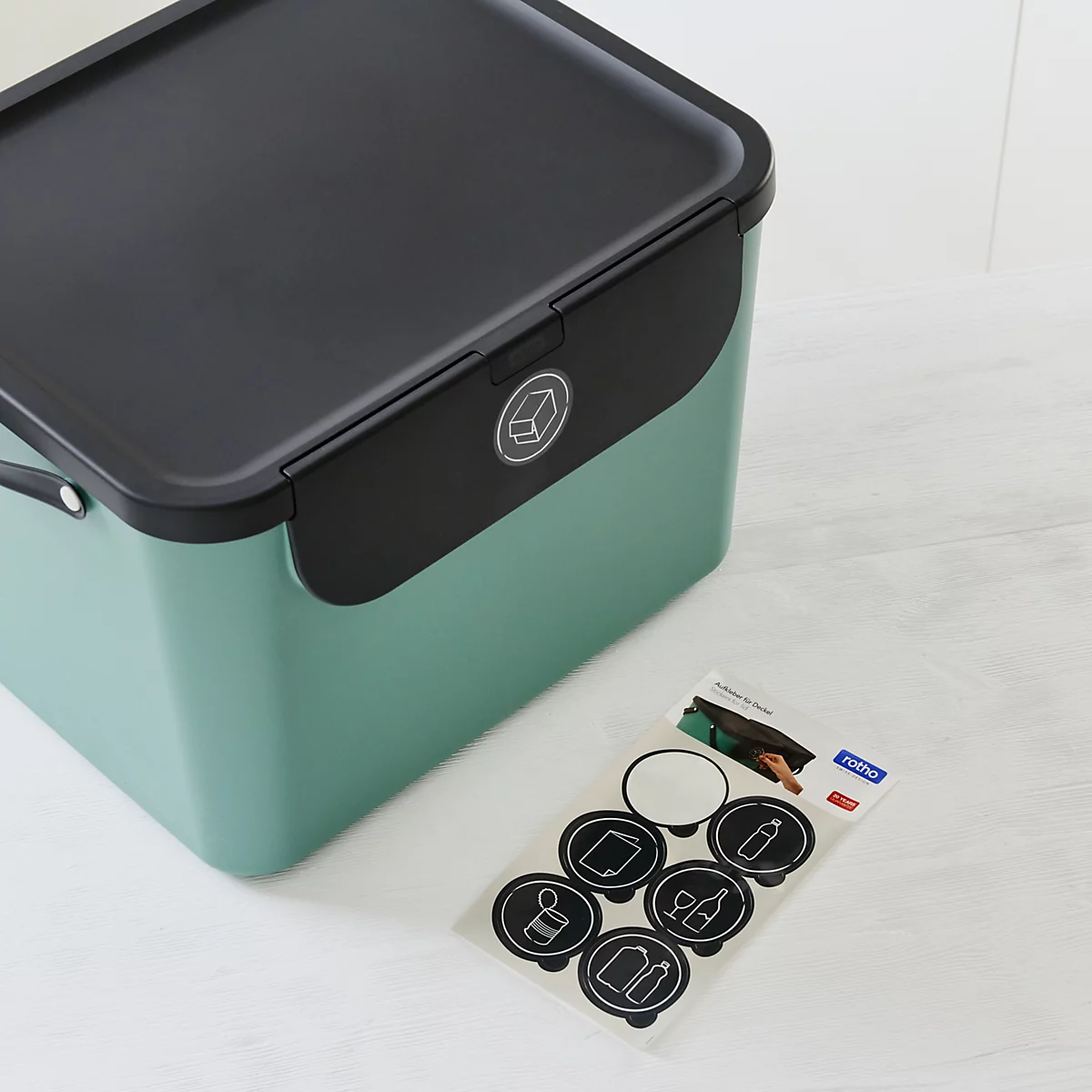 Grüne Mülleimer mit Deckeln. Drei verschieden grosse Behälter stehen vor weisser Wand. Auf den Deckeln sind Symbole abgebildet.