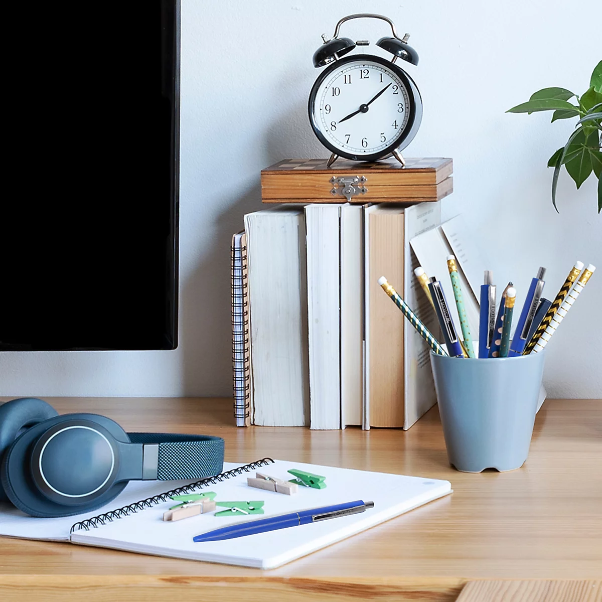 Auf einem Holztisch liegen Notizbuch, Stifte, Kopfhörer und eine Uhr. Die Uhr zeigt kurz vor 9 Uhr.
