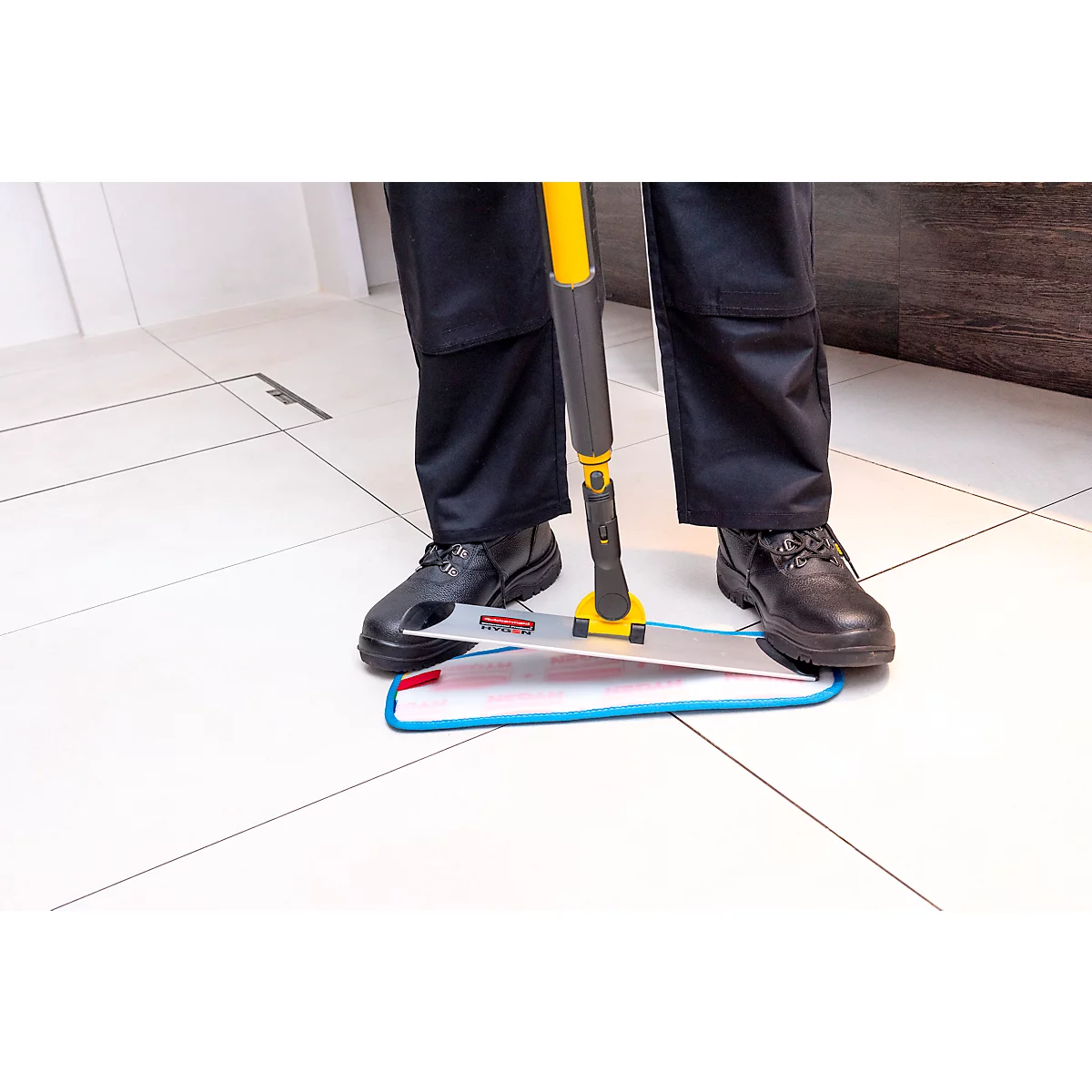 A person stands with a yellow and grey mop on white tiles. The person is wearing black trousers and shoes.