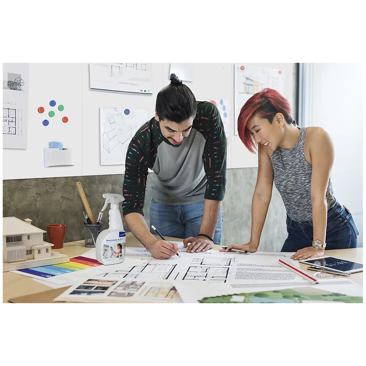 Two people are looking at construction plans. One is drawing, the other is watching. A spray bottle and color samples are on the table.