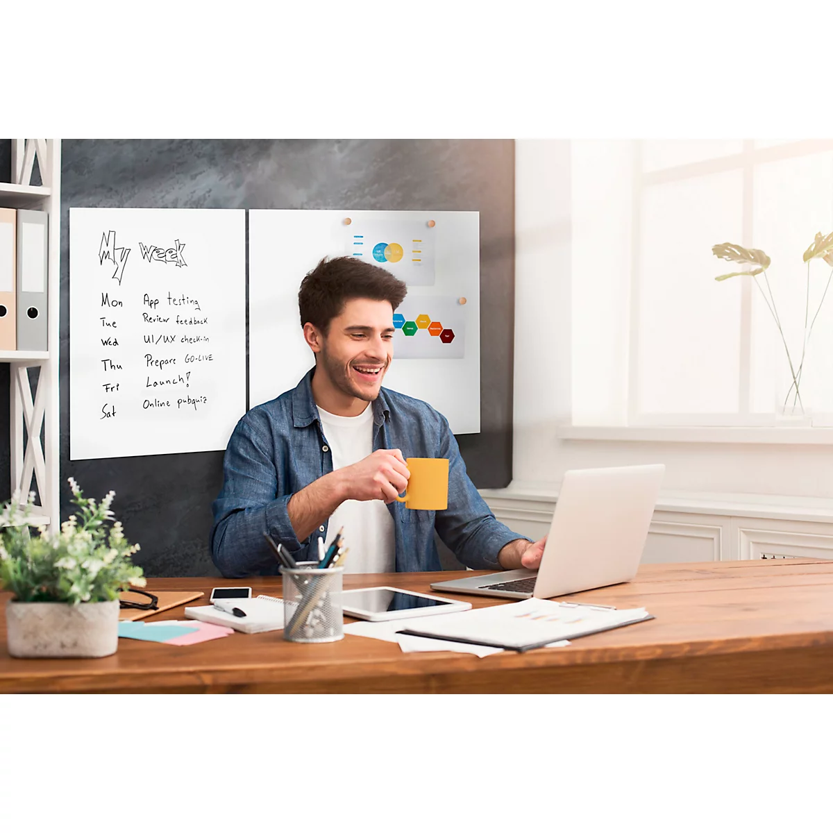 Ein lächelnder Mann sitzt an einem Schreibtisch und arbeitet mit einem Laptop. Er hält eine gelbe Tasse.