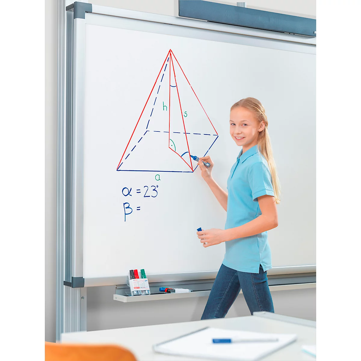 Ein Mädchen zeichnet eine Pyramide auf einer Tafel. Auf der Tafel stehen mathematische Formeln. Sie lächelt in die Kamera.
