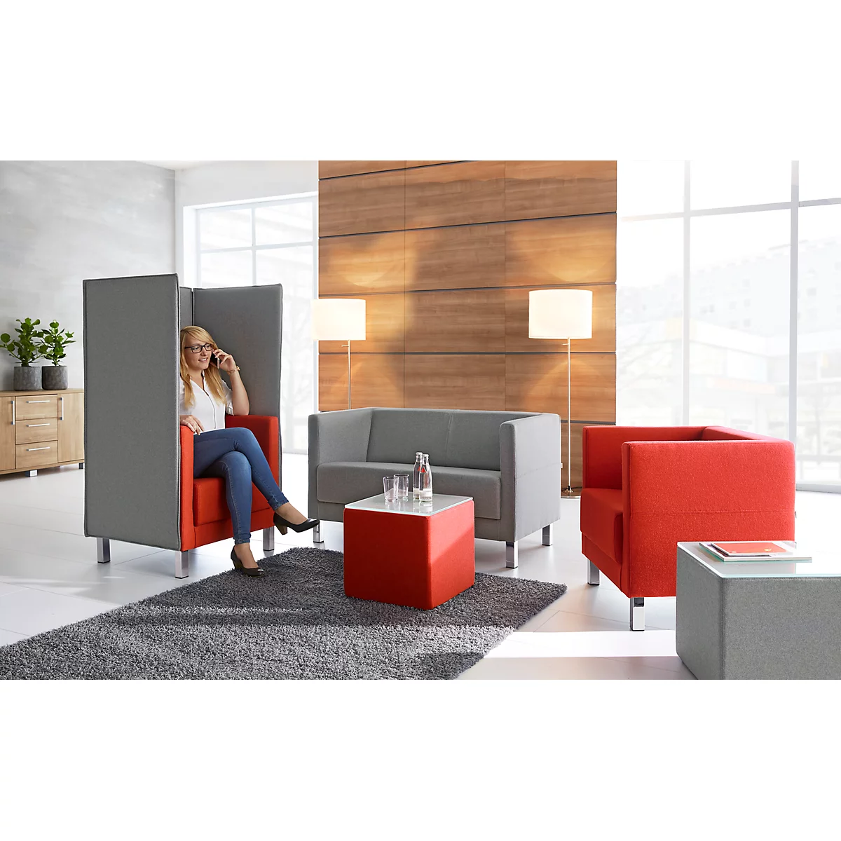 Una mujer hablando por teléfono en un sillón rojo, dentro de un separador de ambientes gris, otros muebles.