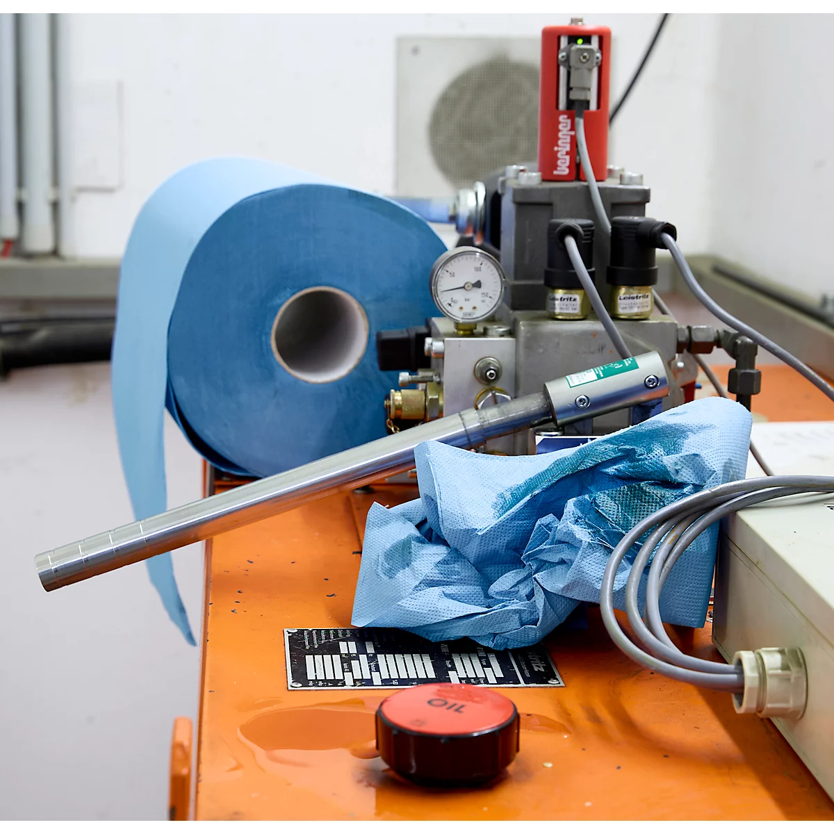 Industrielle Werkbank mit blauer Papierrolle, Messgeräten, Kabeln und einer kleinen, runden roten Öl-Kappe.