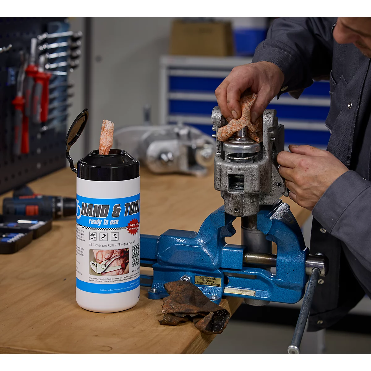 Ein Mann reinigt einen Metallmechanismus an einem Schraubstock. Daneben steht eine Flasche mit Reinigungstüchern.