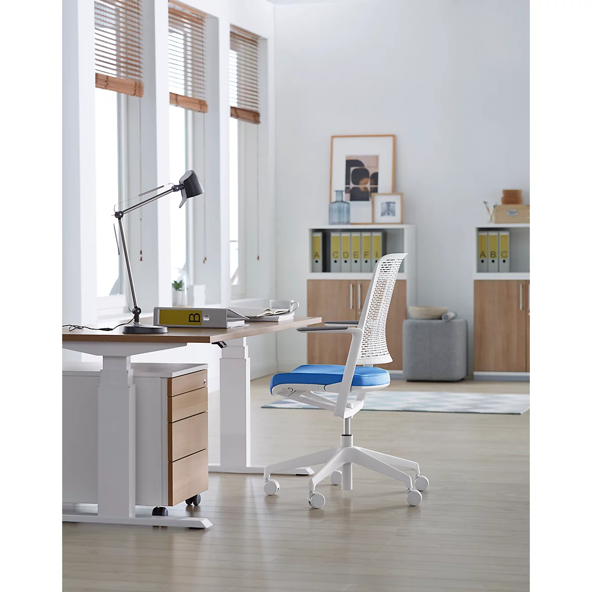 Bright office room with desk and white chair. Windows with blinds and light wooden floor. There is a lamp on the table.