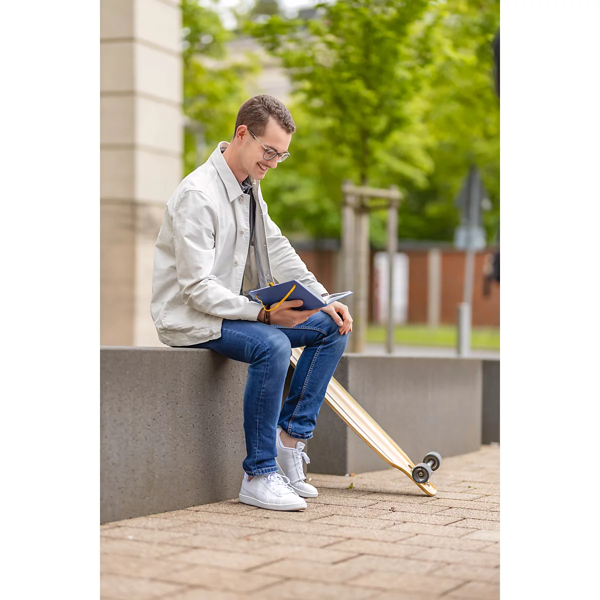Junger Mann mit Brille sitzt auf Mauer, schaut ins Notizbuch, lächelt. Skateboard liegt daneben. Grün im Hintergrund.