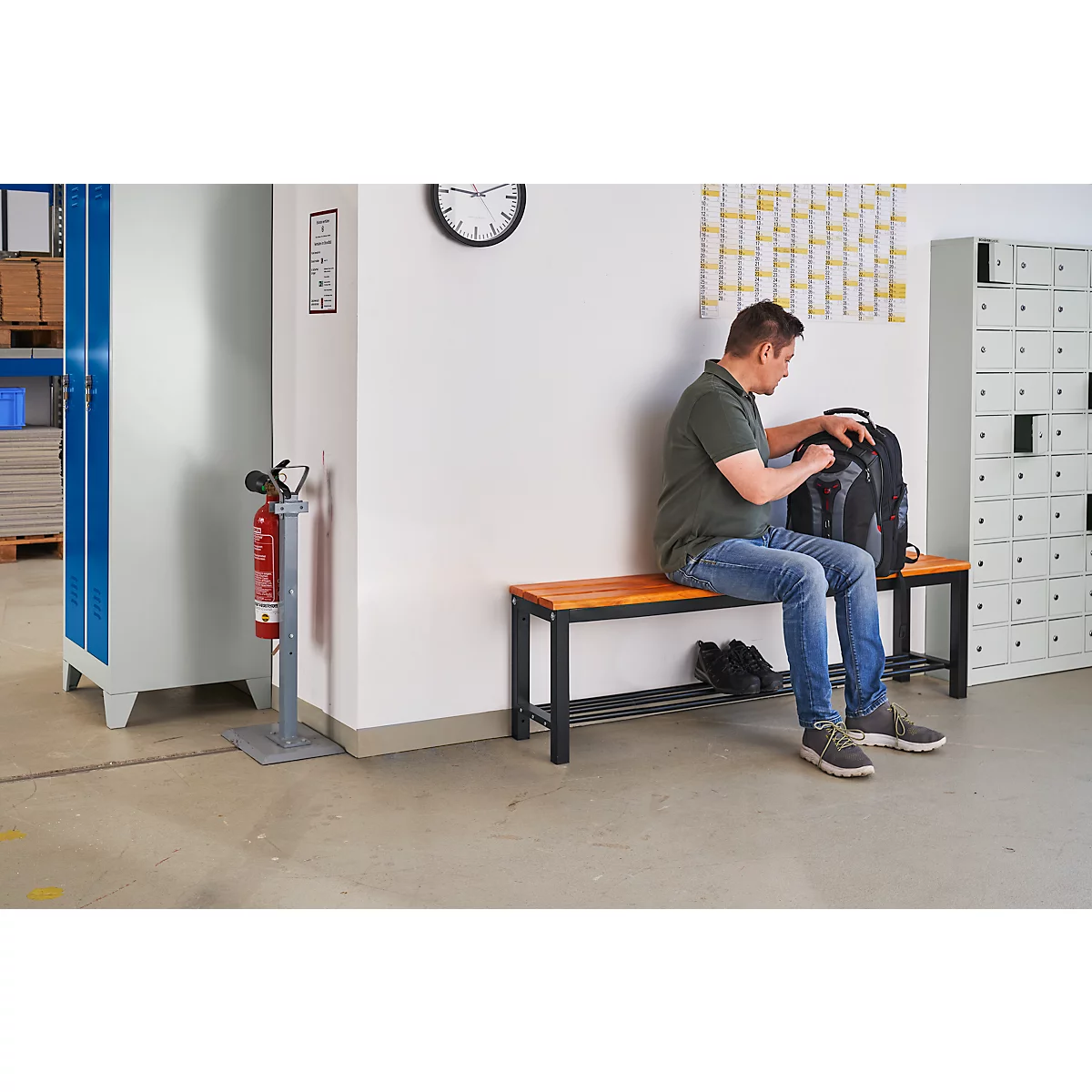 A man sitting on a bench stowing something in his backpack. Shoes are next to him. In the background, a locker and a fire extinguisher.