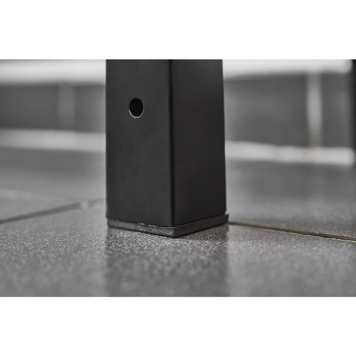 Close-up of a black metal leg with a hole, standing on gray tiled flooring.