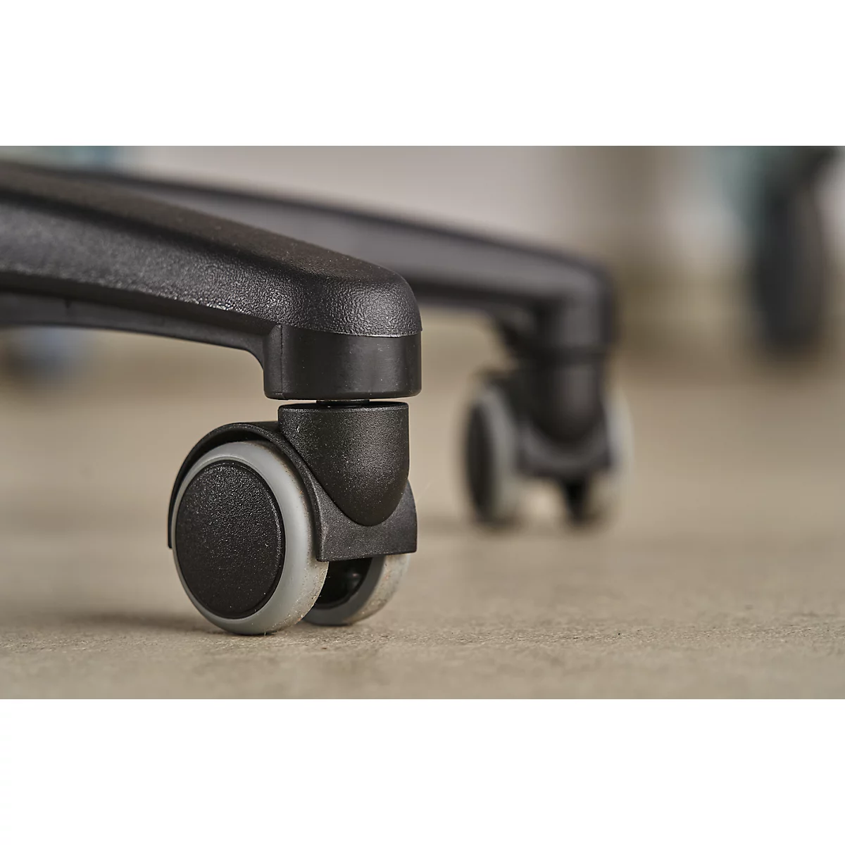Close-up of a black office chair leg with casters. The chair is on a light floor, another chair is blurred in the background.
