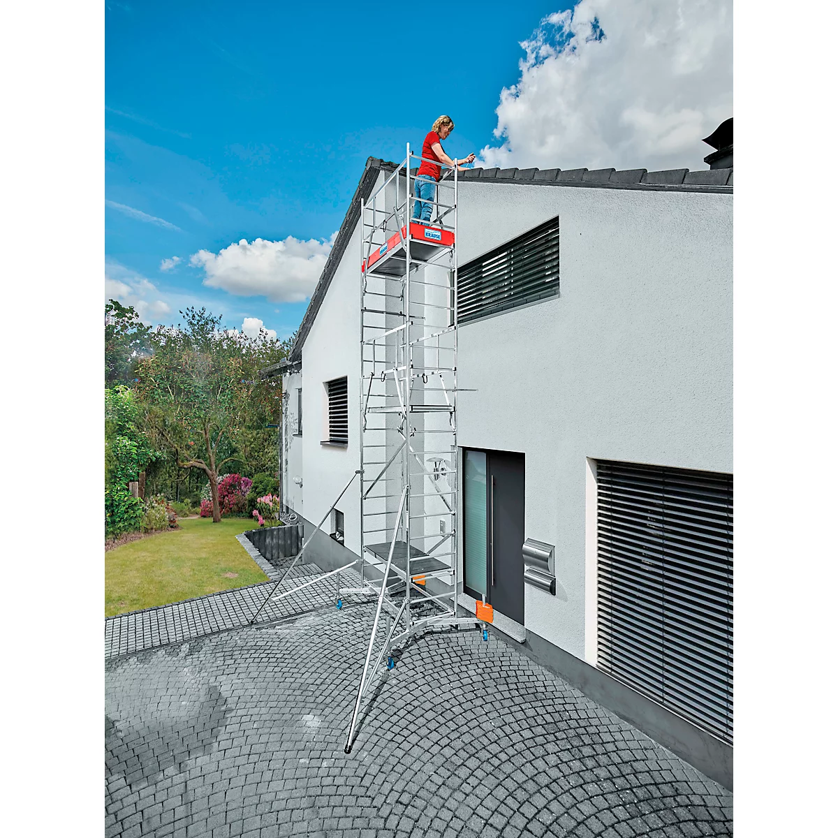 A person stands on scaffolding and works on a house roof. The sky is blue.