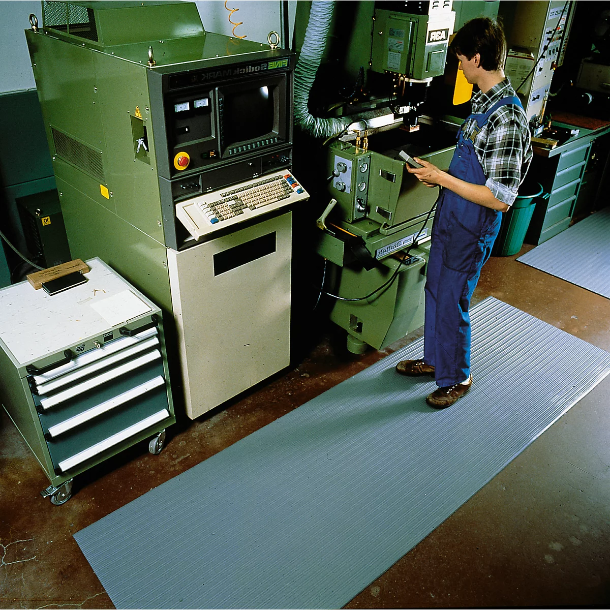 A man in a workshop stands on a gray mat in front of a machine tool. In the background are other machines and tools.
