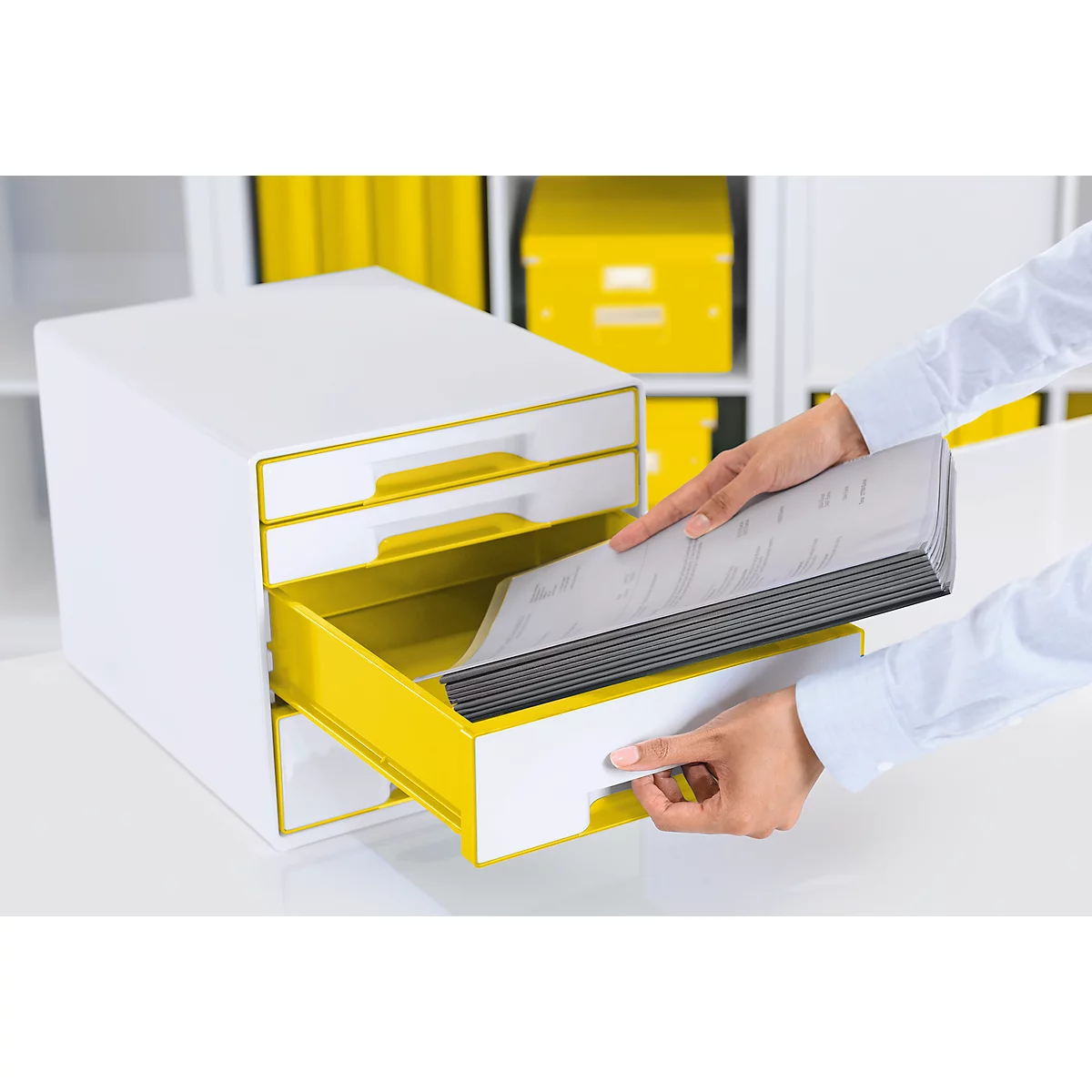Hands placing files in a yellow drawer of a white cabinet. More yellow boxes in the background.