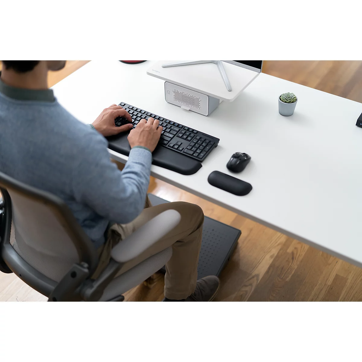 Person typing on keyboard at desk. Mouse and wrist rest next to it. Pot with plant on the table.