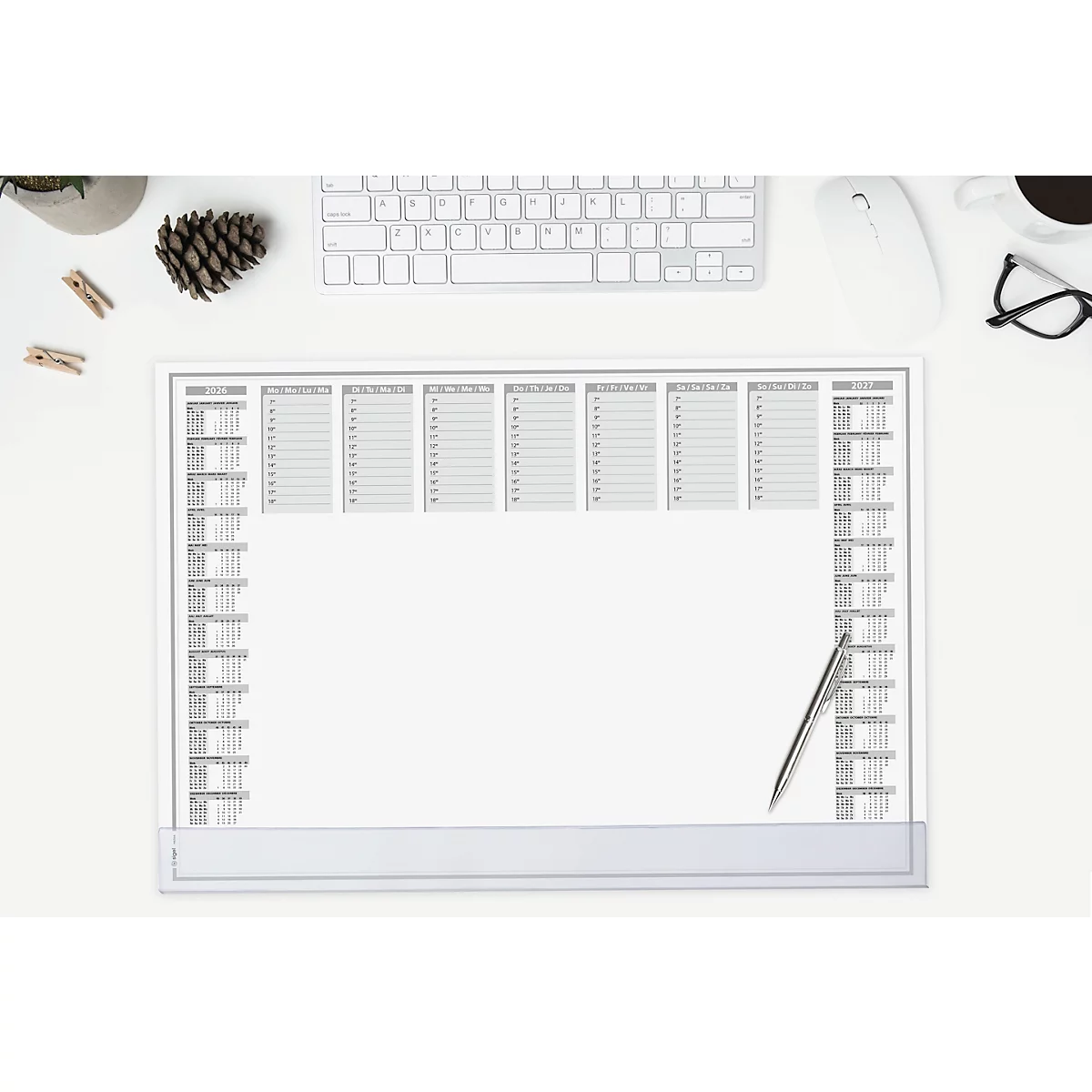 Top view of an empty weekly planner with pen on a white desk. Keyboard, mouse, and accessories are around.