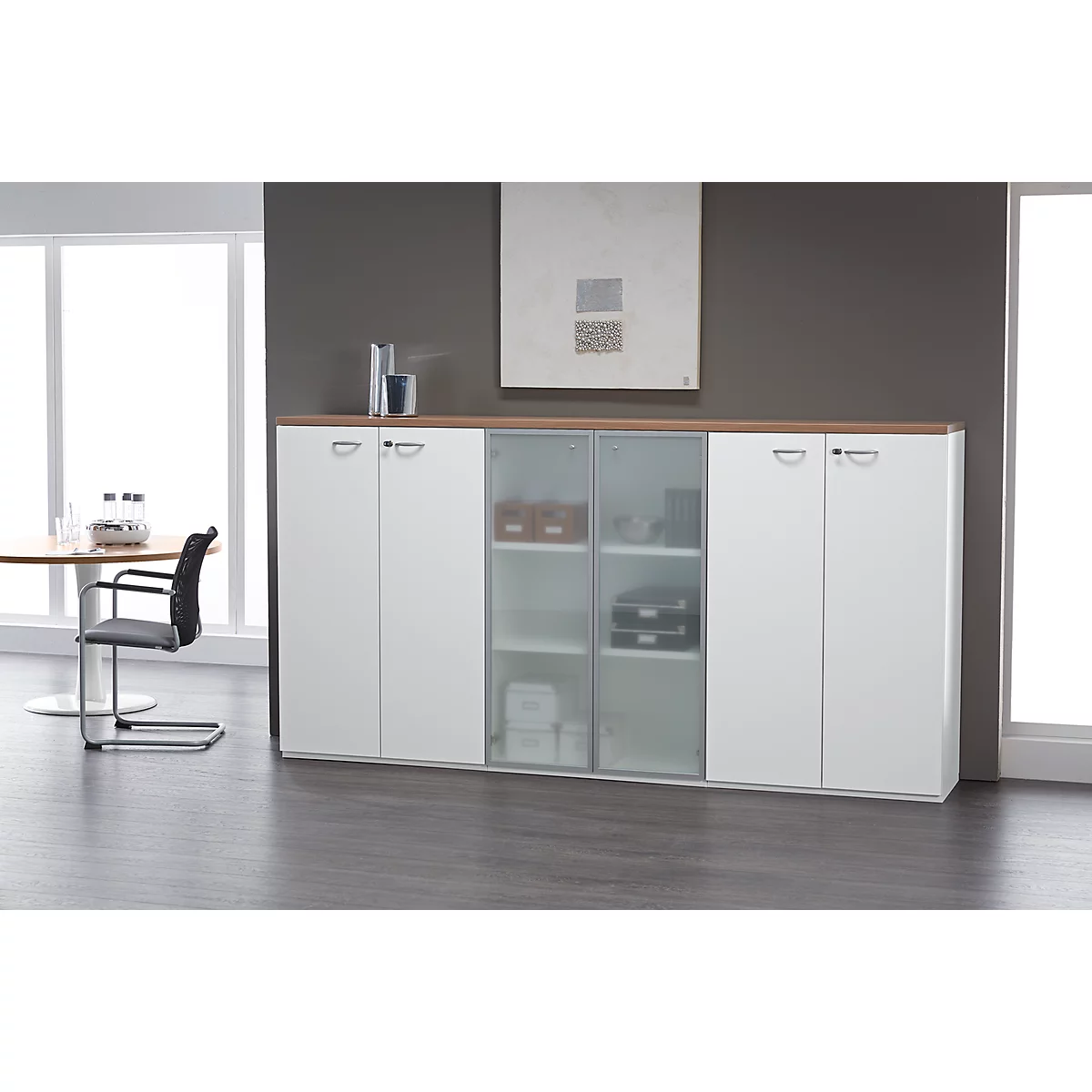 White cabinet with glass and wood doors. A table with chairs in the background. A picture hangs on the wall.