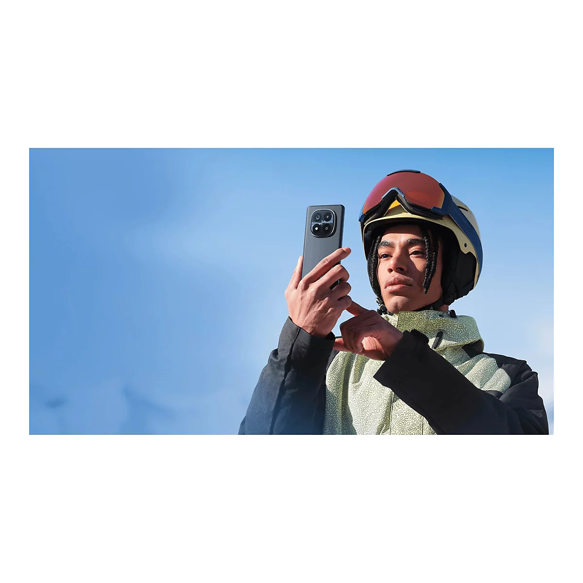 Ein junger Mann mit Skihelm macht ein Foto mit seinem Smartphone. Blauer Himmel im Hintergrund.