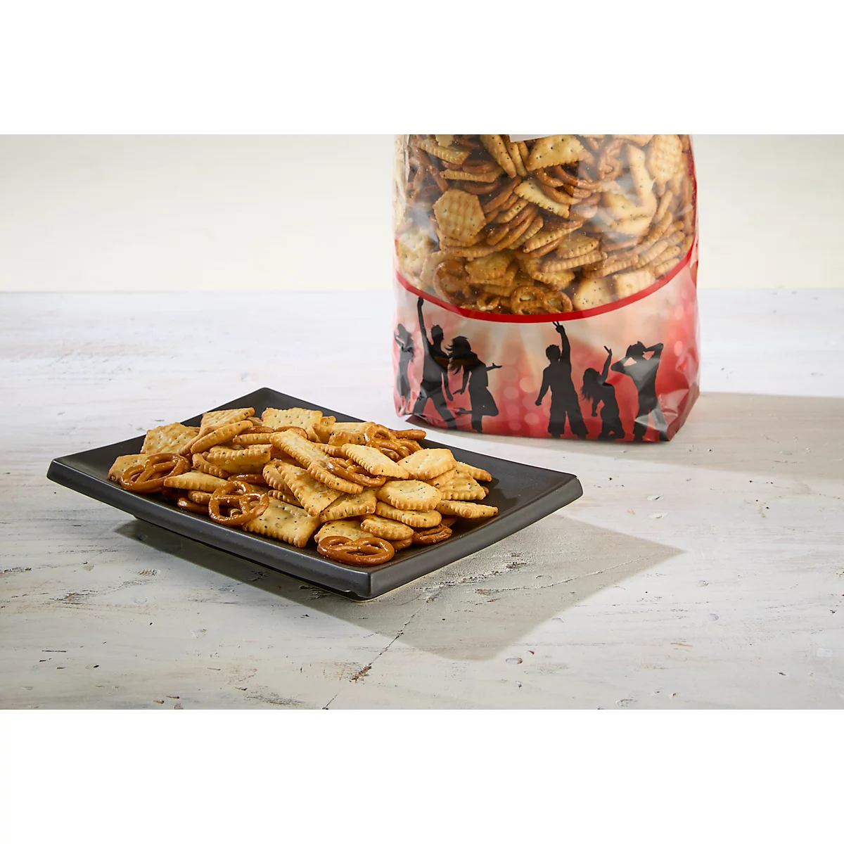 Knabbereien auf einem schwarzen quadratischen Teller, daneben eine Tüte voller Cracker. Auf der Tüte sind tanzende Silhouetten abgebildet.