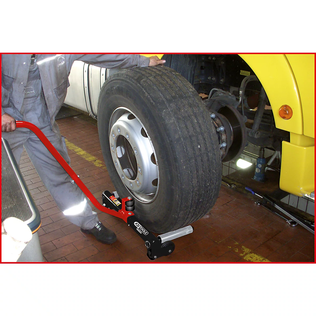 Ein Mechaniker montiert mit einem roten Werkzeug einen Reifen an einem gelben LKW. Reifen und Werkzeug sind im Detail zu sehen.