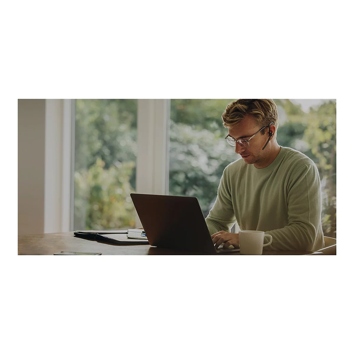 Ein Mann mit Brille und Headset arbeitet am Laptop. Auf dem Tisch liegen Unterlagen, ein Becher Kaffee steht daneben. Im Hintergrund verschwommene Bäume.