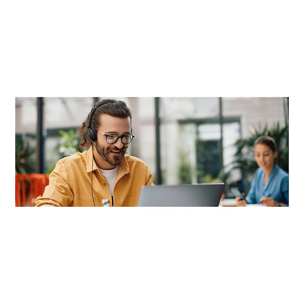 Ein Mann mit Brille und Headset arbeitet am Laptop, lÃ¤chelt. Im Hintergrund eine Frau.