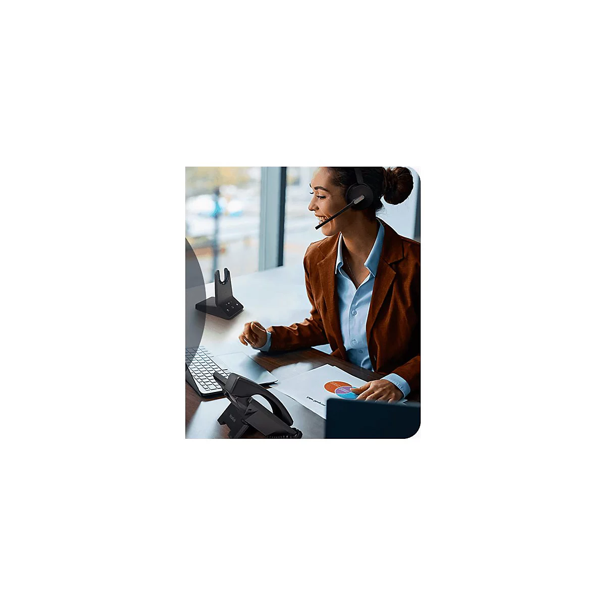 Eine Frau mit Headset arbeitet am Schreibtisch. Sie betrachtet Diagramme und einen Laptop.