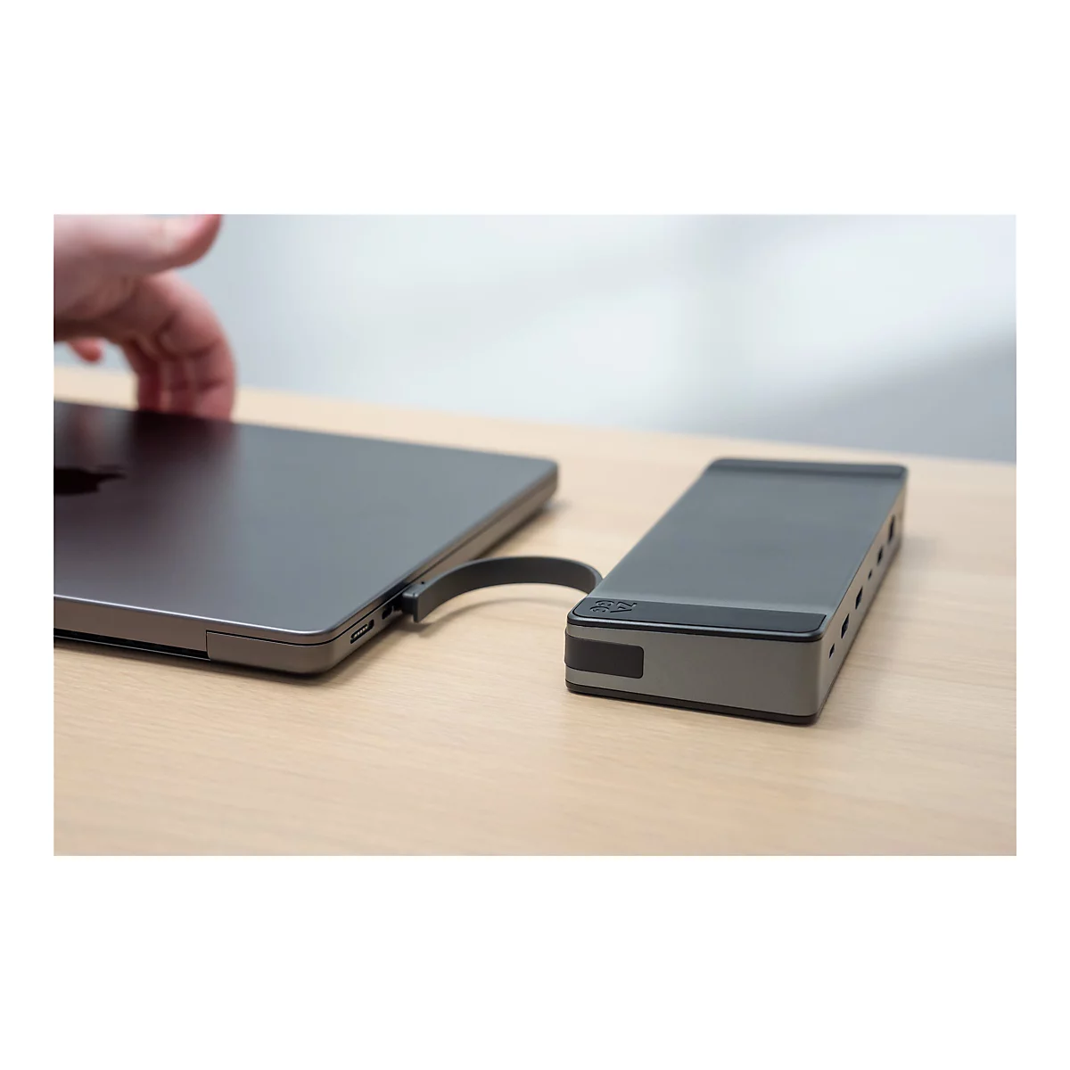 Un ordinateur portable gris et un hub gris sont reliés par un câble, sur une table en bois clair. Une main s'approche de l'ordinateur portable.